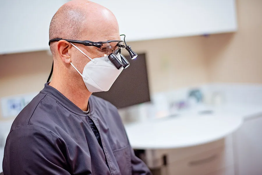 A dentist wearing a face mask and surgical loupes with a headlight in a dental office.